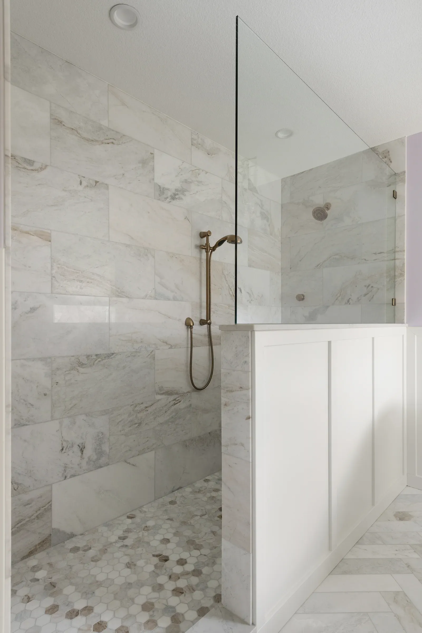 Walk-in shower with large-format marble tile, brass handheld fixture, mosaic floor, and fixed glass panel – primary bathroom remodel in Elko New Market MN by Honey-Doers Remodeling