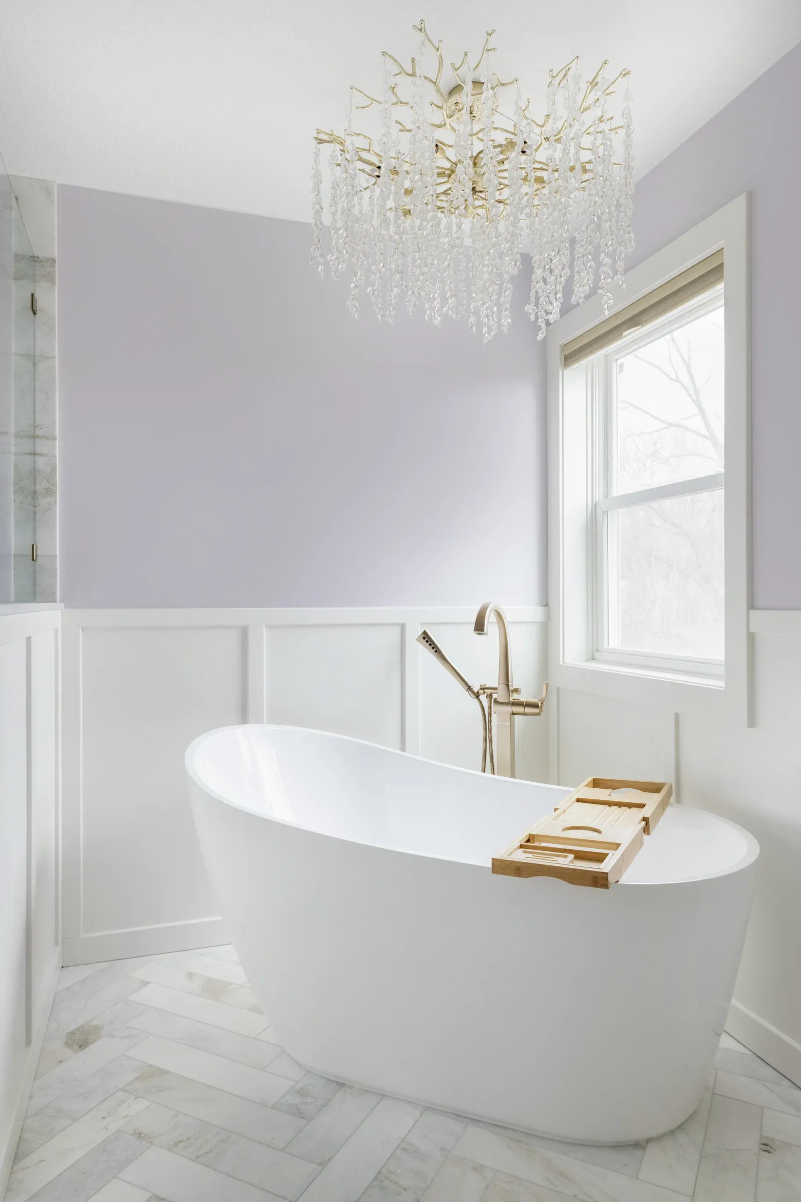 Freestanding soaking tub with crystal chandelier overhead, lavender accent wall, white wainscoting, and herringbone marble floor – Elko New Market primary bathroom remodel by Honey-Doers Remodeling