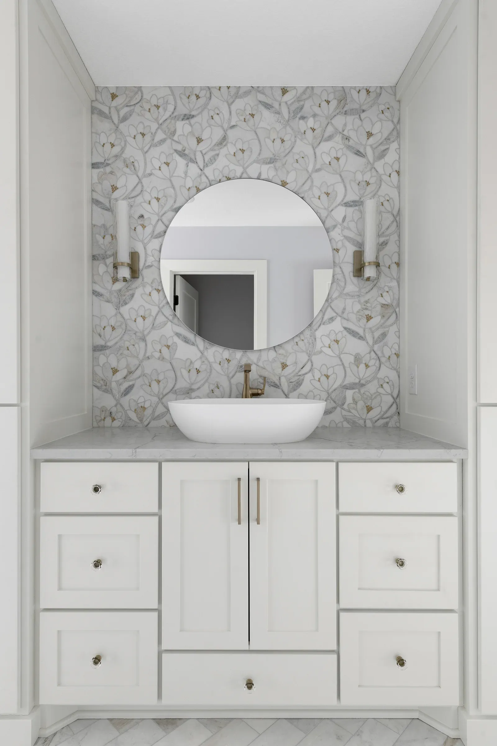 Vanity niche with floral mosaic tile accent wall, round mirror, brass wall sconces, vessel sink, and white shaker cabinetry – Elko New Market primary bathroom by Honey-Doers Remodeling