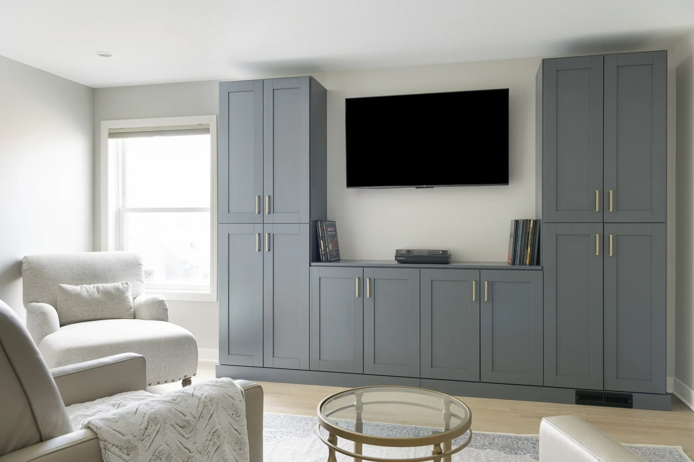 Close view of blue-gray semi-custom cabinet wall with wall-mounted TV, brass hardware, and hardwood floors – home office and entertainment remodel in Elko New Market MN by Honey-Doers Remodeling