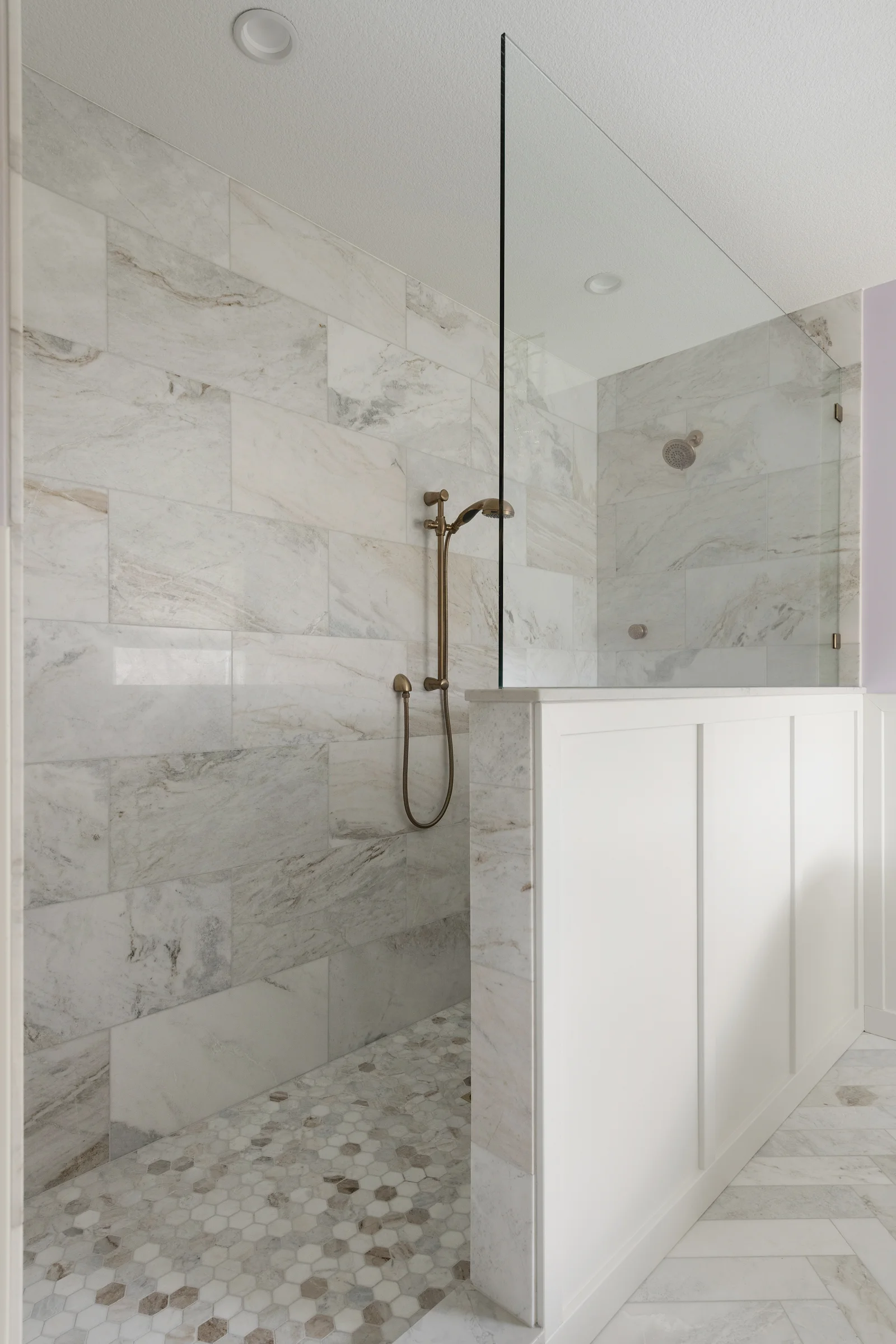 Walk-in shower with large-format marble tile, brass handheld fixture, mosaic floor, and fixed glass panel – primary bathroom remodel in Elko New Market MN by Honey-Doers Remodeling