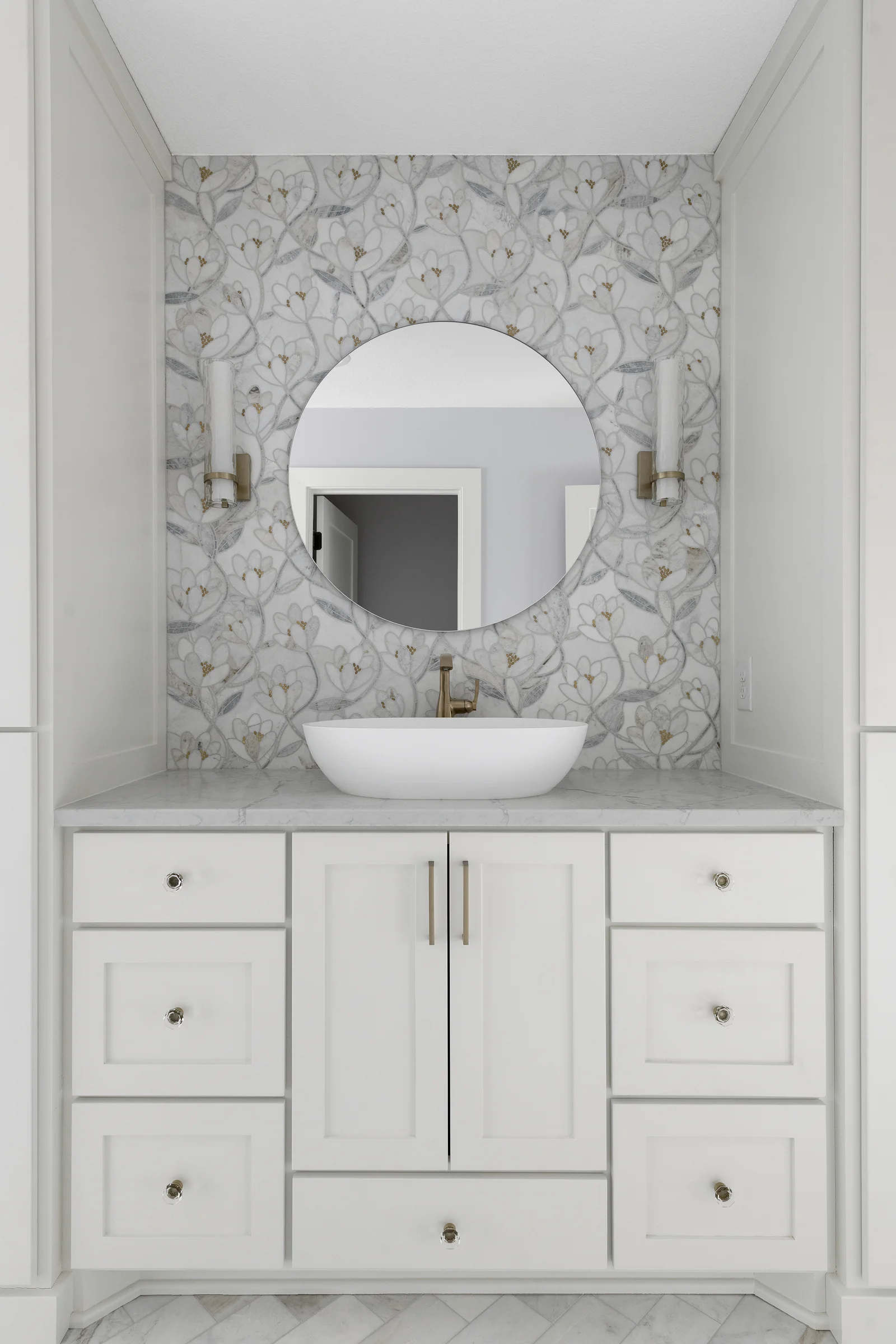 Vanity niche with floral mosaic tile accent wall, round mirror, brass wall sconces, vessel sink, and white shaker cabinetry – Elko New Market primary bathroom by Honey-Doers Remodeling